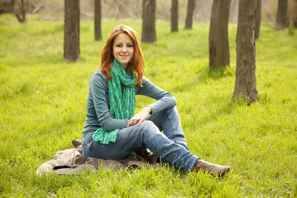 Vacker rödhårig flicka sitter på grönt gräs på park. — Stockfoto