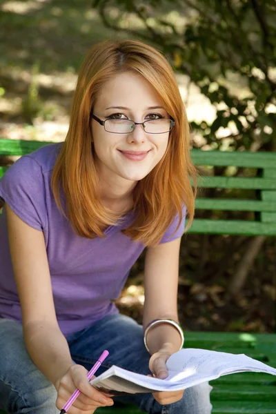 Рыжая девушка в очках делает домашнее задание в парке . — стоковое фото