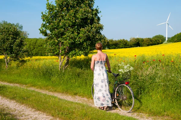 Ελκυστική νεαρή γυναίκα με ένα ποδήλατο παύσεις στην άκρη ενός πεδίου αγριογογγύλης ή/και Φωτογραφία Αρχείου