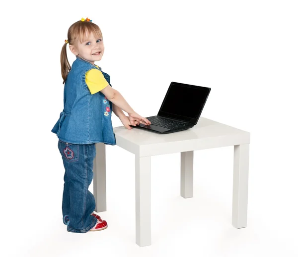 Little girl in blue jeans standing at a table with a laptop — Stock Photo, Image