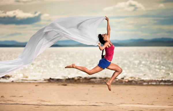 Beautiful young woman jumping on the beach with a white tissue — Zdjęcie stockowe