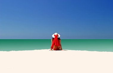 Woman in Red hat and bikini sitting all alone on empty beach clipart