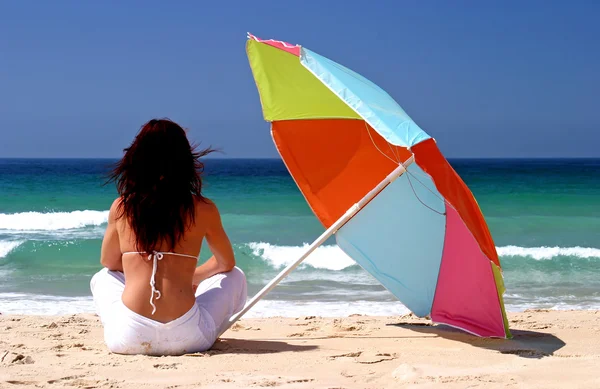 Woman sitting under colorful parasol on white sandy beach — Stock Photo, Image