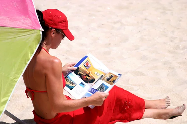 Młoda kobieta w czerwonym bikini, siedząc na plaży, czytanie magazin — Zdjęcie stockowe