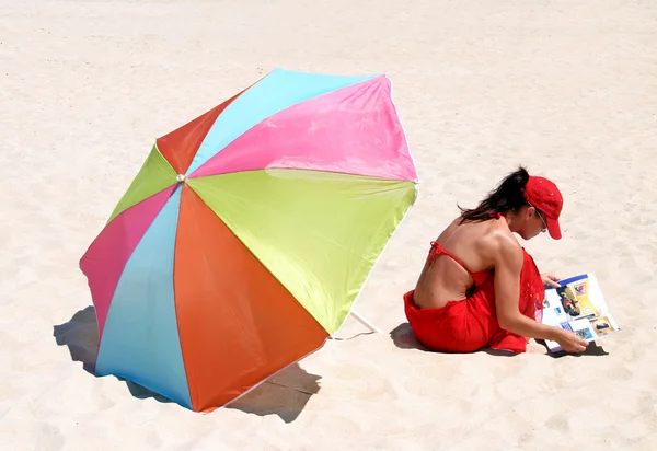 Kırmızı bikinili kumsalda magazin okuma oturan genç kadın — Stok fotoğraf
