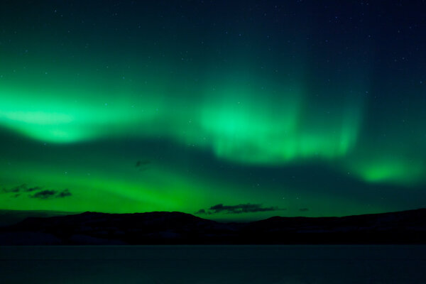 Green northern lights (aurora borealis) substorm above silhouette of hills and clouds.