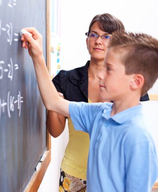 Boy writing on chalkboard in front of teacher clipart