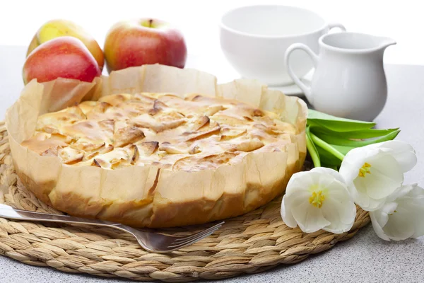 Apple pie, tulips, apples and glassware on a wicker mat — Stock Photo, Image