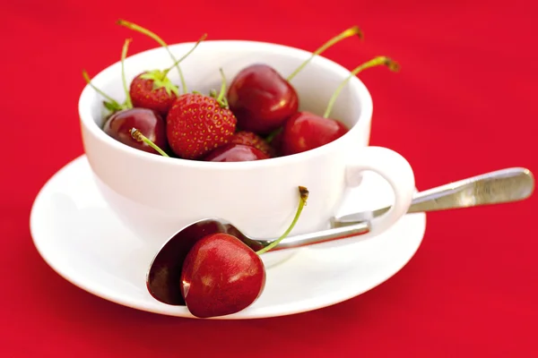 Taza platillo cuchara cereza y fresa sobre un fondo rojo —  Fotos de Stock