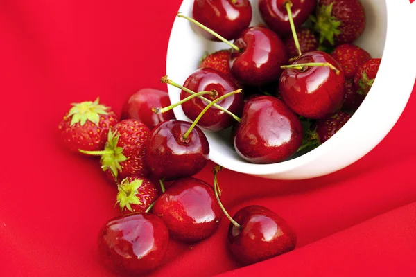 Cerezas y fresas en una taza blanca sobre un fondo rojo —  Fotos de Stock