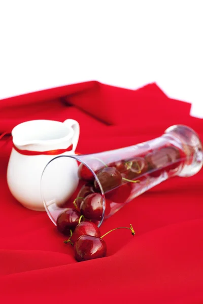Молочный кувшин с ленточным стаканом вишни на красном фоне — стоковое фото