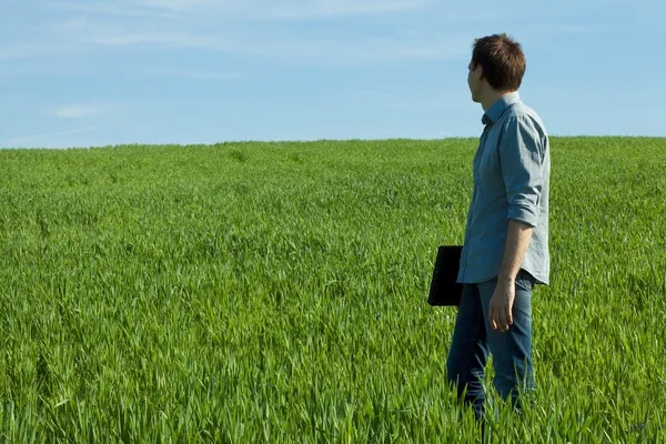 Молодой человек стоит с ноутбуком в зеленом поле — стоковое фото