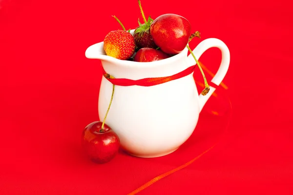 Mjölk kannan band körsbär och jordgubbar på en röd bakgrund — Stockfoto