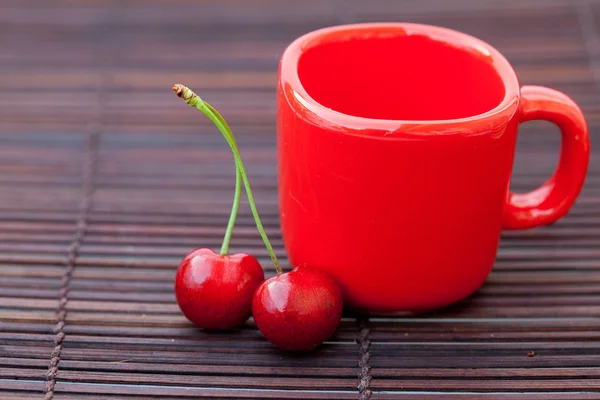Две вишни и красная чашка на бамбуковом коврике. — стоковое фото