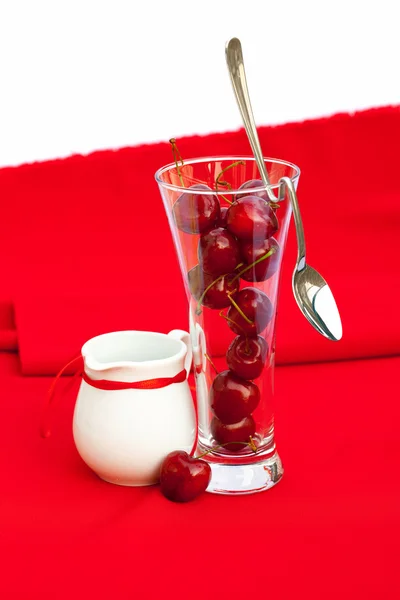 Milchkanne mit Schleife, Glas Kirsche und Löffel auf rotem Rücken — Stockfoto