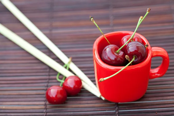 Cherry ,cup and chopsticks on bamboo mat — Stock Photo, Image