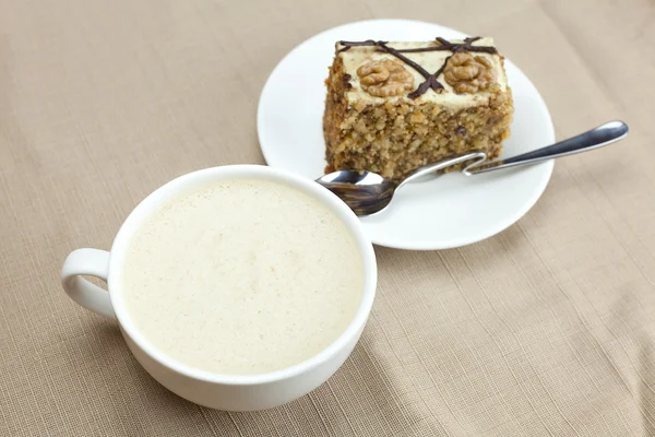 A cup of cappuccino, a piece of cake with nuts and spoon lying o — Stock Photo, Image