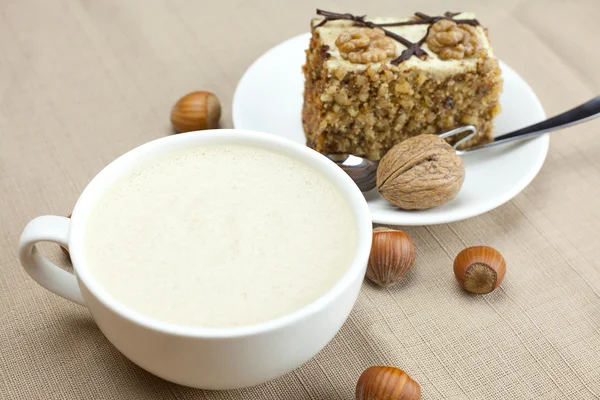 A cup of cappuccino, a piece of cake with nuts and spoon lying o — Stock Photo, Image