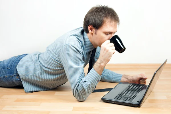 Hombre joven con taza usando el ordenador portátil acostado en el suelo en la habitación —  Fotos de Stock