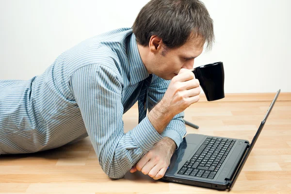 Hombre joven con taza usando el ordenador portátil acostado en el suelo en la habitación — Foto de Stock
