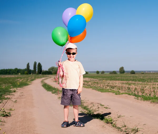 Little boy with colorful balloons standing on the country road — Stock Photo, Image