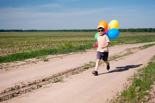 Little boy with colorful balloons running by the country road — Stock Photo, Image