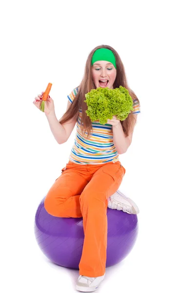 Молодая женщина на мяче ест салат и морковь — стоковое фото
