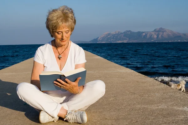 Ανώτερος γυναίκα που κάθεται στην παραλία ανάγνωση βιβλίου στις καλοκαιρινές διακοπές στη Μαγιόρκα — Φωτογραφία Αρχείου
