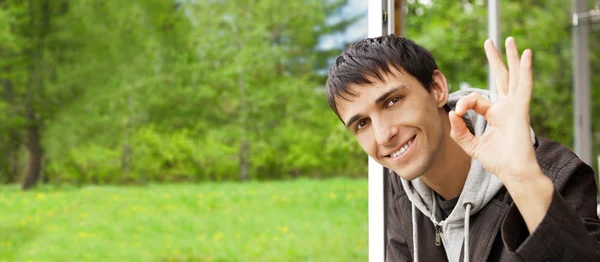 Young brunette man gives the OK sign, standing near big window a — Stock Photo, Image