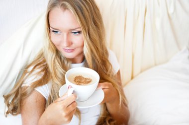 Smiling woman drinking a coffee lying on a bed at home or hotel. clipart