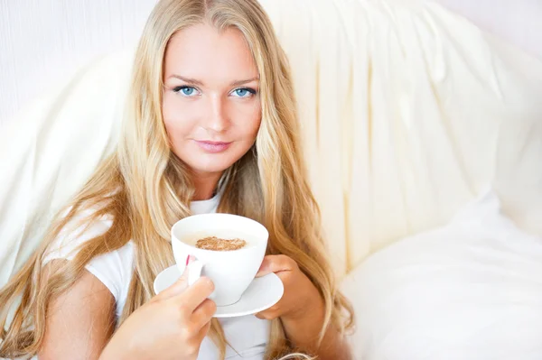 Lachende vrouw drinken een kopje koffie liggend op een bed thuis of hotel. — Stockfoto