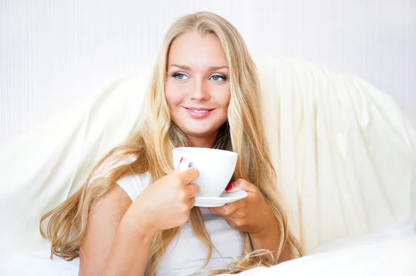 Mujer sonriente bebiendo un café acostada en una cama en casa o en un hotel . — Foto de Stock