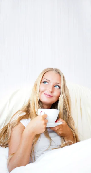 Mujer sonriente bebiendo un café acostada en una cama en casa o en un hotel . —  Fotos de Stock
