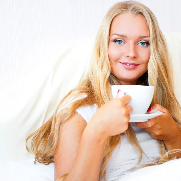 Donna sorridente che beve un caffè sdraiata su un letto a casa o in hotel . — Foto Stock