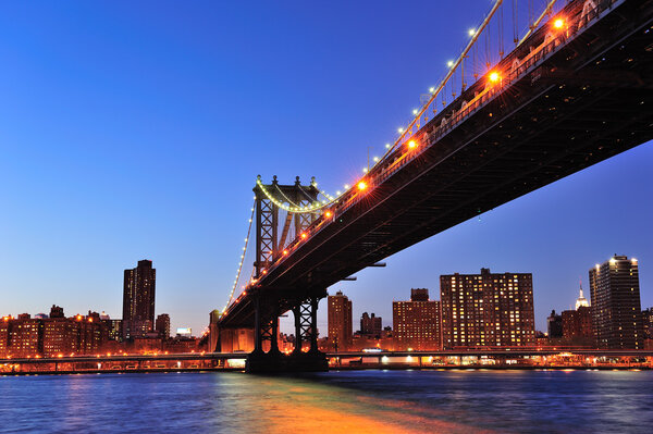 New York City Manhattan Bridge over East River at dusk illuminated with light with reflections and downtown skyline viewed from Brooklyn.
