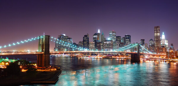 New York City Brooklyn Bridge and Manhattan skyline panorama view with skyscrapers over Hudson River illuminated with lights at dusk after sunset.