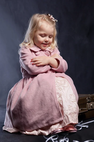 Chica rizada en abrigo rosa se sienta en la maleta de edad —  Fotos de Stock