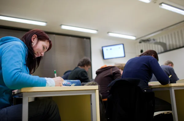 Hübsche Studentin sitzt bei einer Prüfung in einem Klassenzimmer voller — Stockfoto