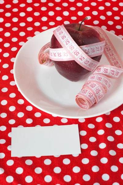 Червоне яблуко та вимірювальна стрічка на білій тарілці на червоному тлі та картці — стокове фото