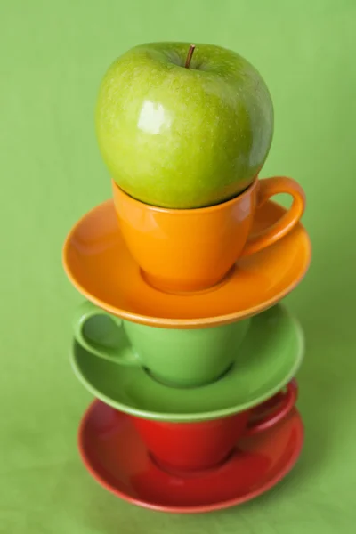 Green apple and bright cups on a green background — Stock Photo, Image