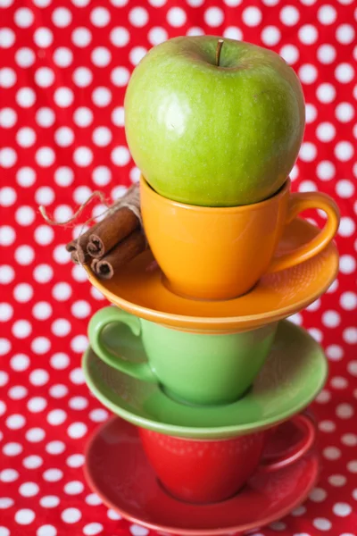 Green apple, cinnamon sticks and bright cups with a red background — Stock Photo, Image