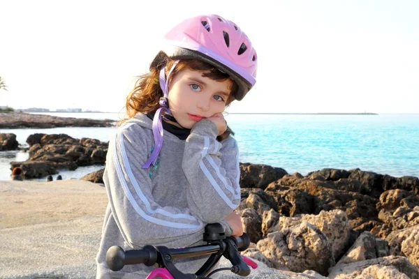 Bicicletta piccola ragazza pensosa casco rosa in spiaggia rocciosa — Foto Stock