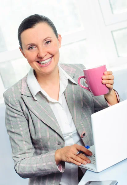 Sonriente joven mujer de negocios con taza usando el ordenador portátil —  Fotos de Stock