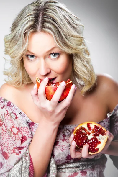 Blonde woman biting pomegranate on gray — Stock Photo, Image
