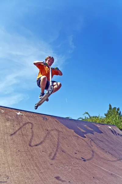 Menino está pulando com uma scooter sobre uma coluna vertebral no parc skate e — Fotografia de Stock
