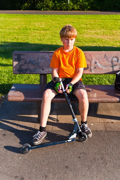 Jongen zittend op een bankje met zijn scooter — Stockfoto