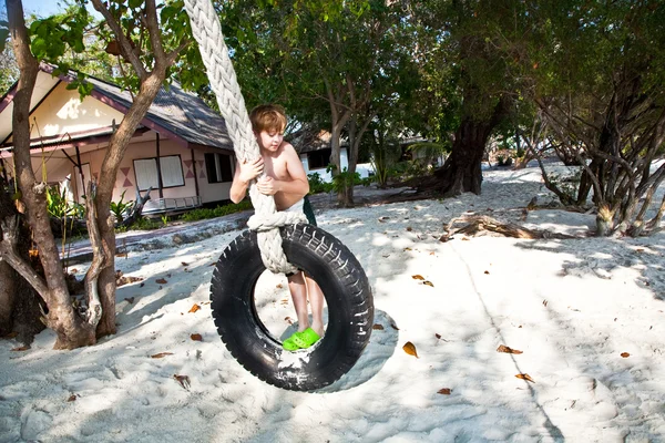Menino sentado em um balanço feito de um pneu velho na praia sob tr — Fotografia de Stock