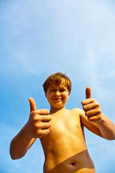 Glada leende ung pojke med bakgrunden blå himmel ger fingersig — Stockfoto