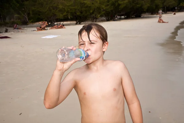 Young happy boy with brown wet hair is drinking water at the bea — Stock Photo, Image