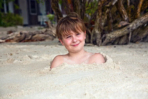 Счастливый мальчик, покрытый мелким песком на пляже — стоковое фото